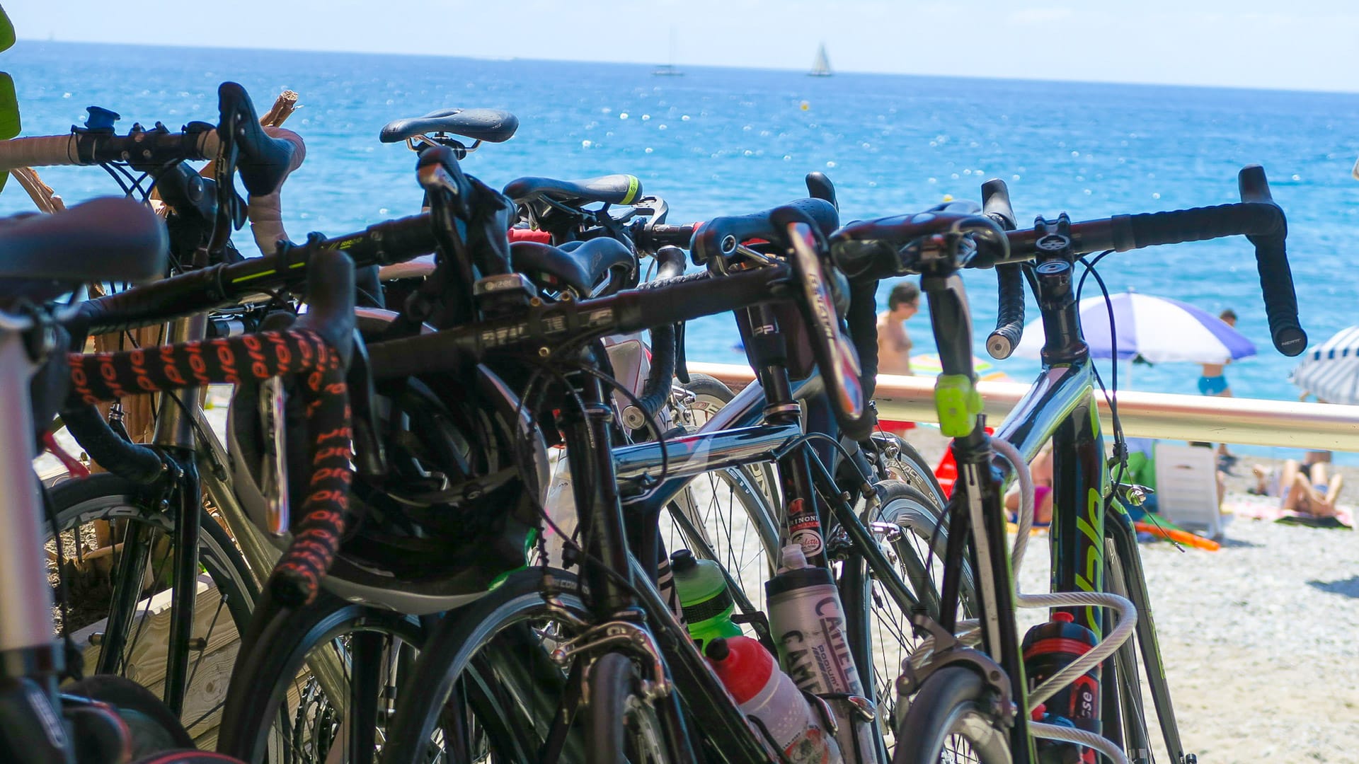 Road bikes lined up by a beach with people sunbathing and sailing in the background