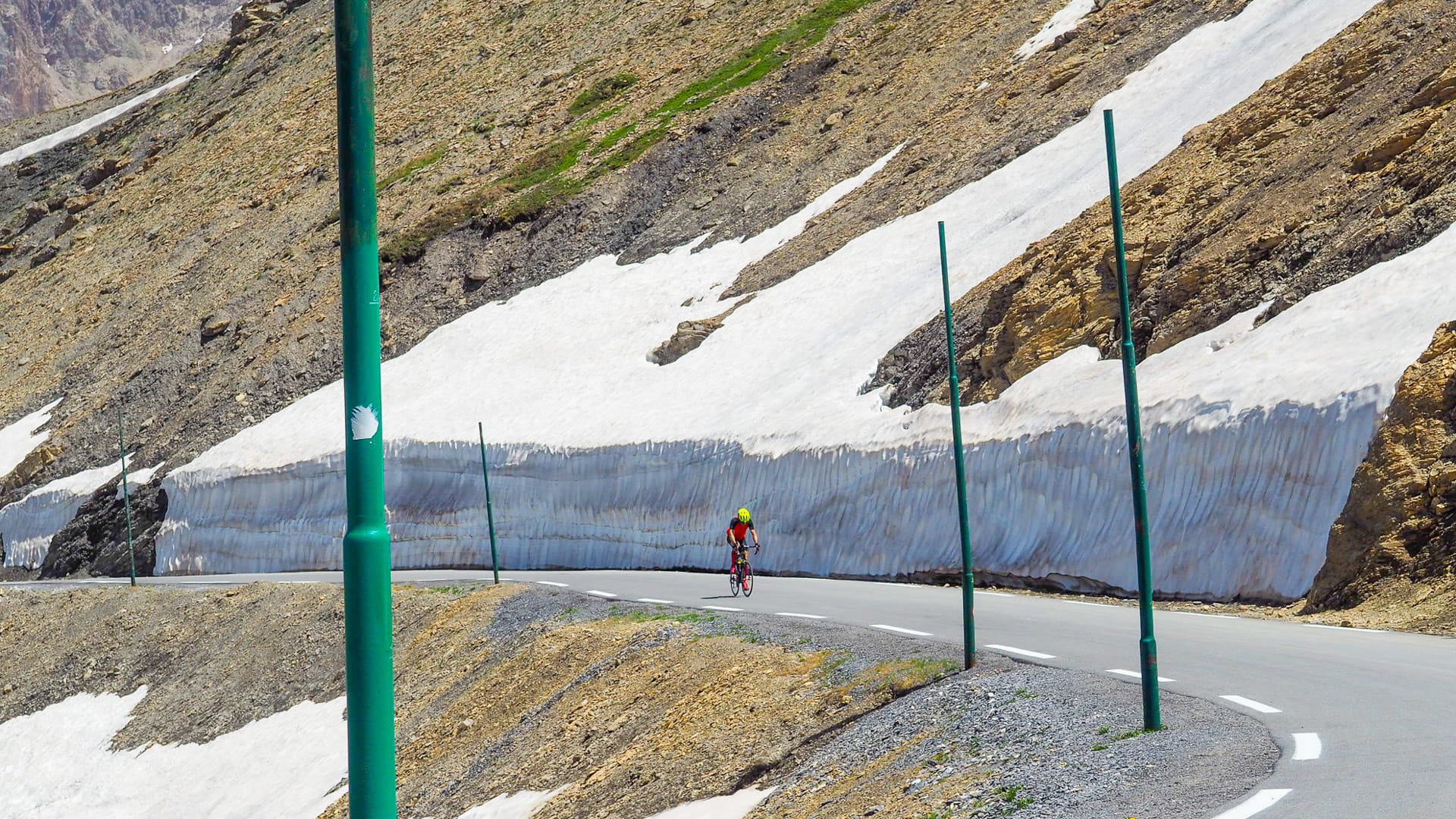 Cyclist riding past snowbanks on high alpine pass road with green guide poles on the Route des Grandes Alpes
