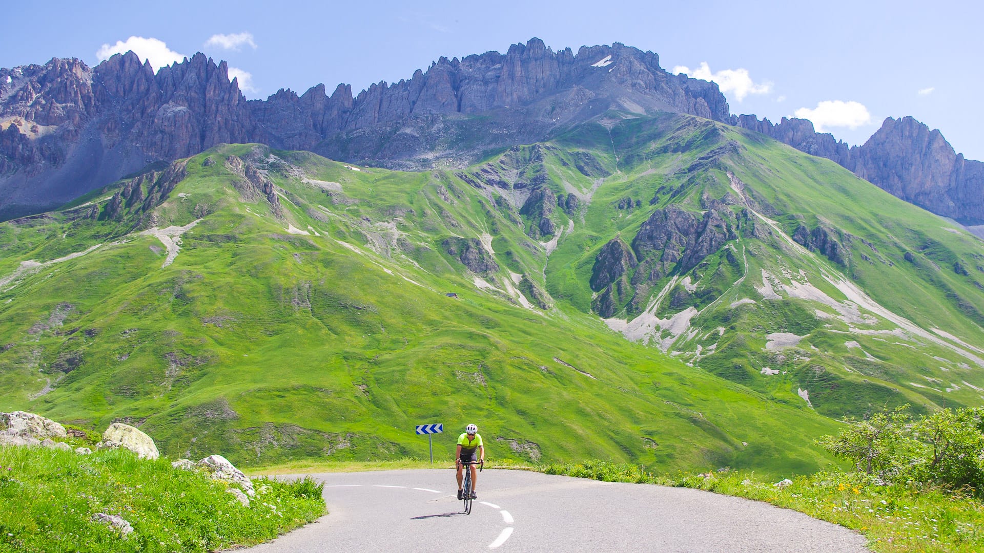 Solo cyclist descending green alpine valley below jagged peaks on the Route des Grandes Alpes, France