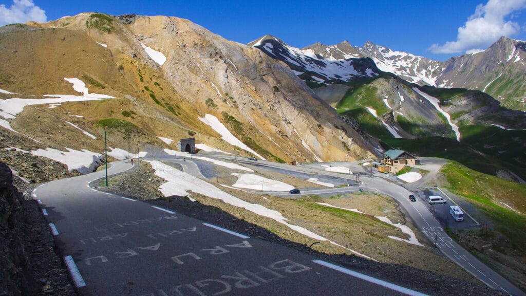 Cyclist climbing steep hairpins of Col du Galibier on the Route des Grandes Alpes, with snow patches and peaks