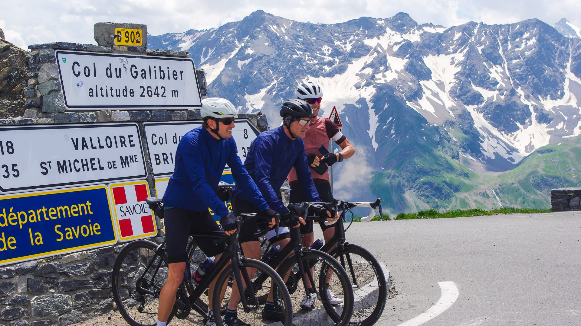 Cyclists stopped at Col du Galibier summit sign on Route des Grandes Alpes with snow-covered peaks in the background