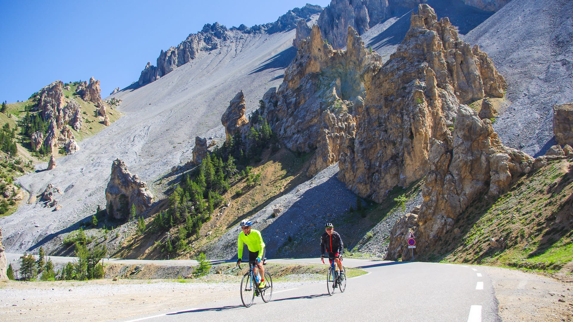 Two cyclists riding past dramatic rock formations on a bright alpine day on the Route des Grandes Alpes
