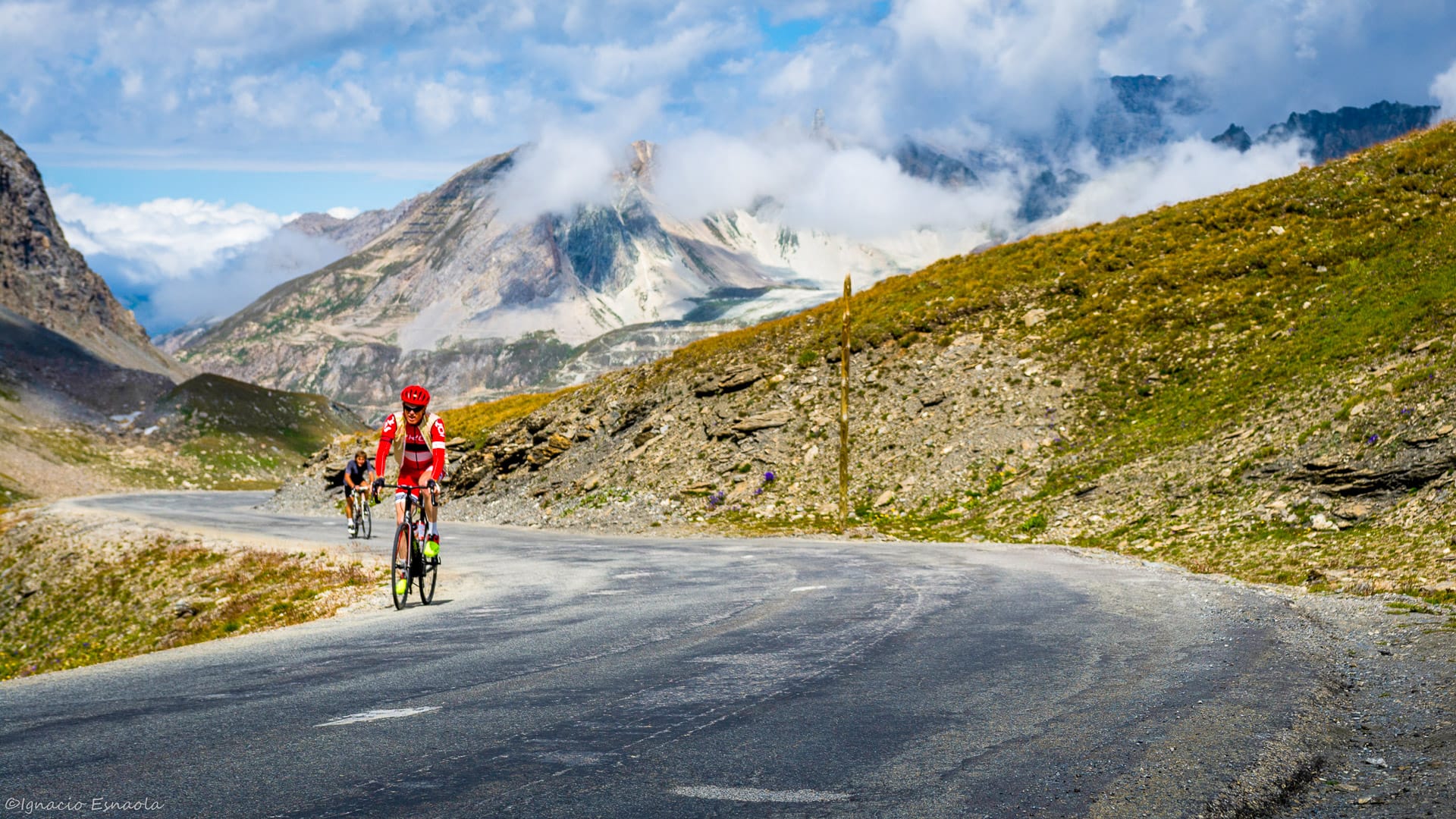 Cyclists climbing dramatic mountain road on the Route des Grandes Alpes under shifting clouds and rocky peaks