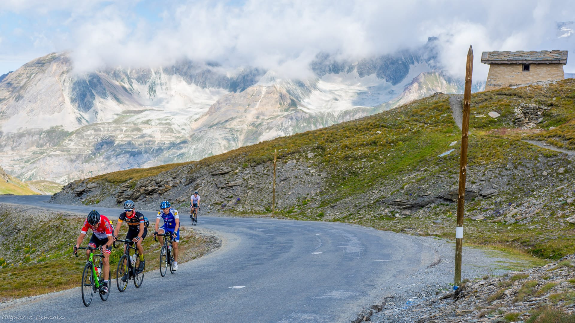 Cyclists climbing high alpine road on Route des Grandes Alpes with dramatic glacial peaks and clouds in the background