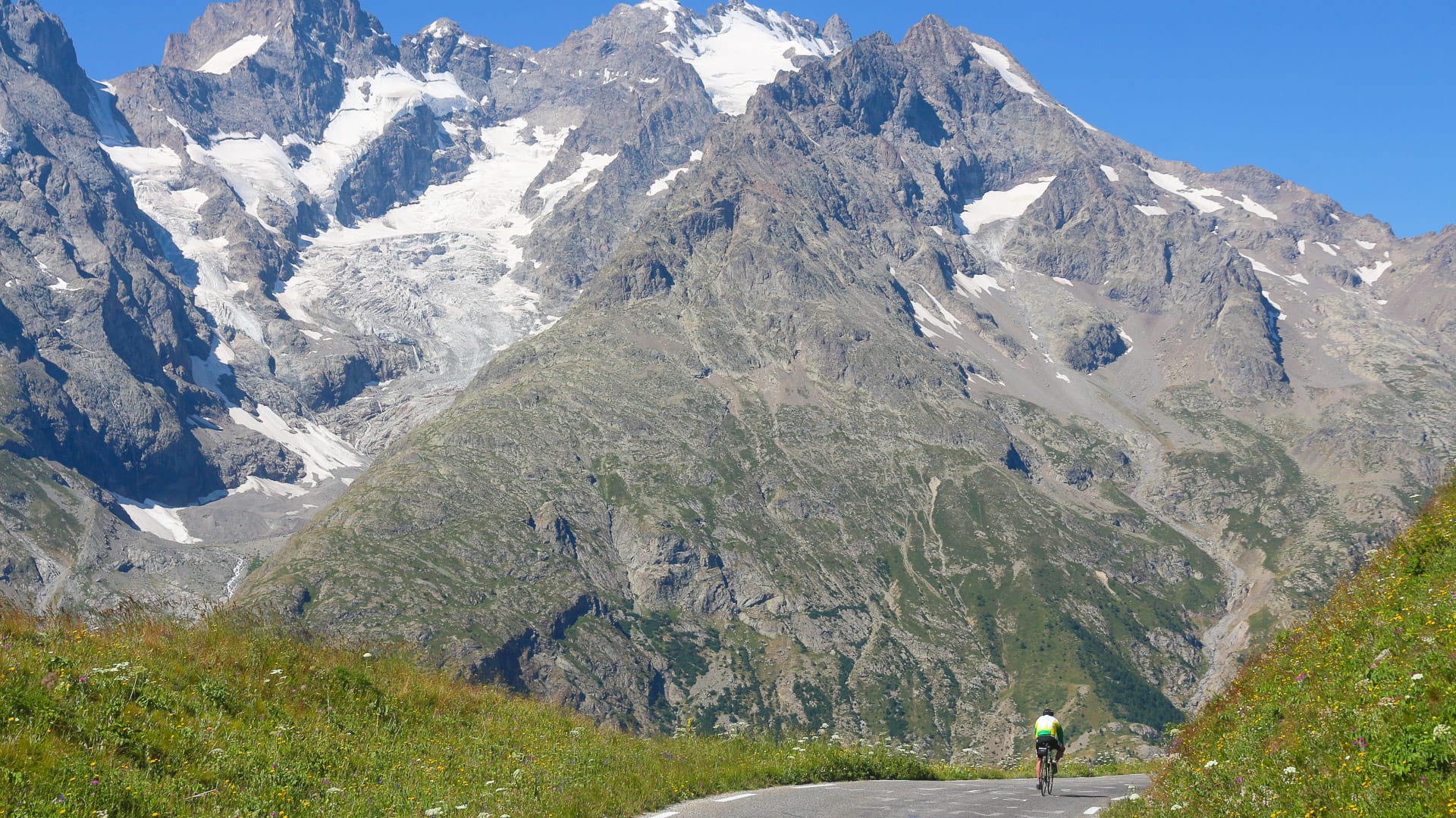 Cyclist climbing beneath towering glaciers on the Route des Grandes Alpes under clear blue skies