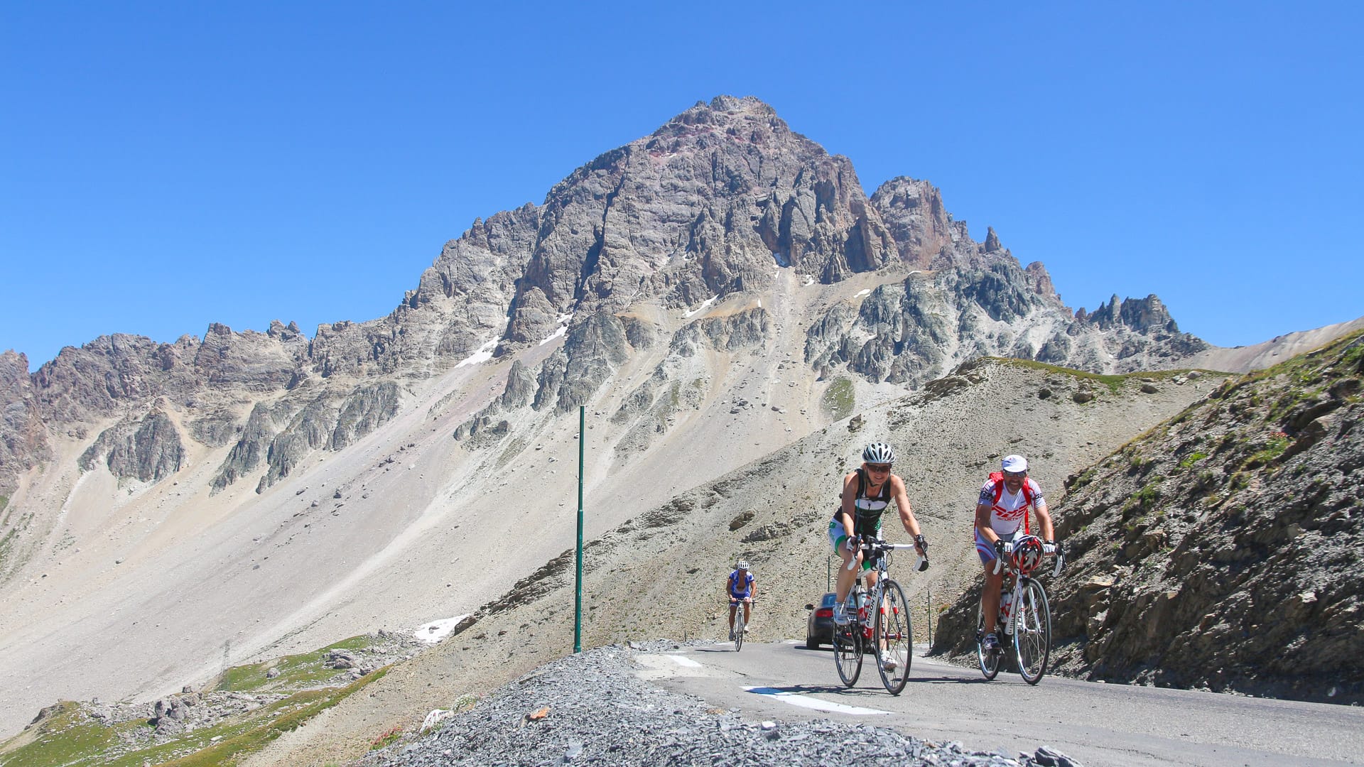 Group of cyclists climbing steep alpine pass beneath rugged rocky peak on the Route des Grandes Alpes
