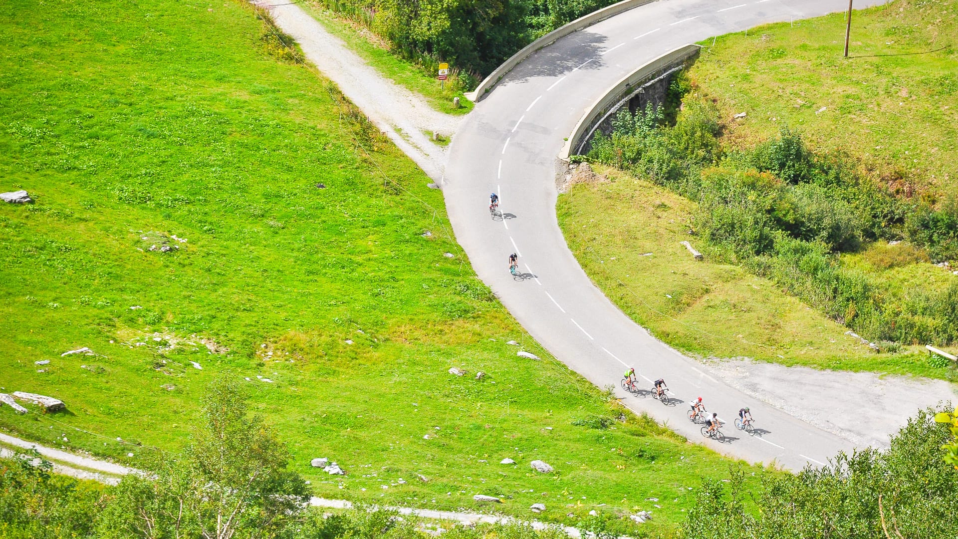 Cyclists descending sweeping bend through green pastures on Route des Grandes Alpes, France