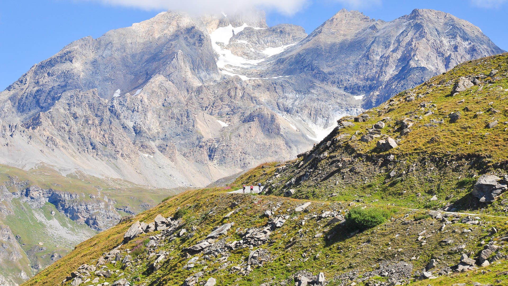 Distant group of cyclists climbing scenic alpine road beneath massive rocky mountain on the Route des Grandes Alpes