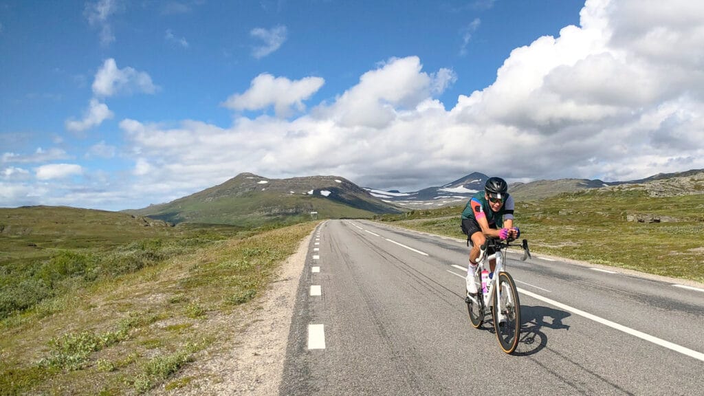 Cyclist riding on a long open road through a mountainous landscape under a partly cloudy sky