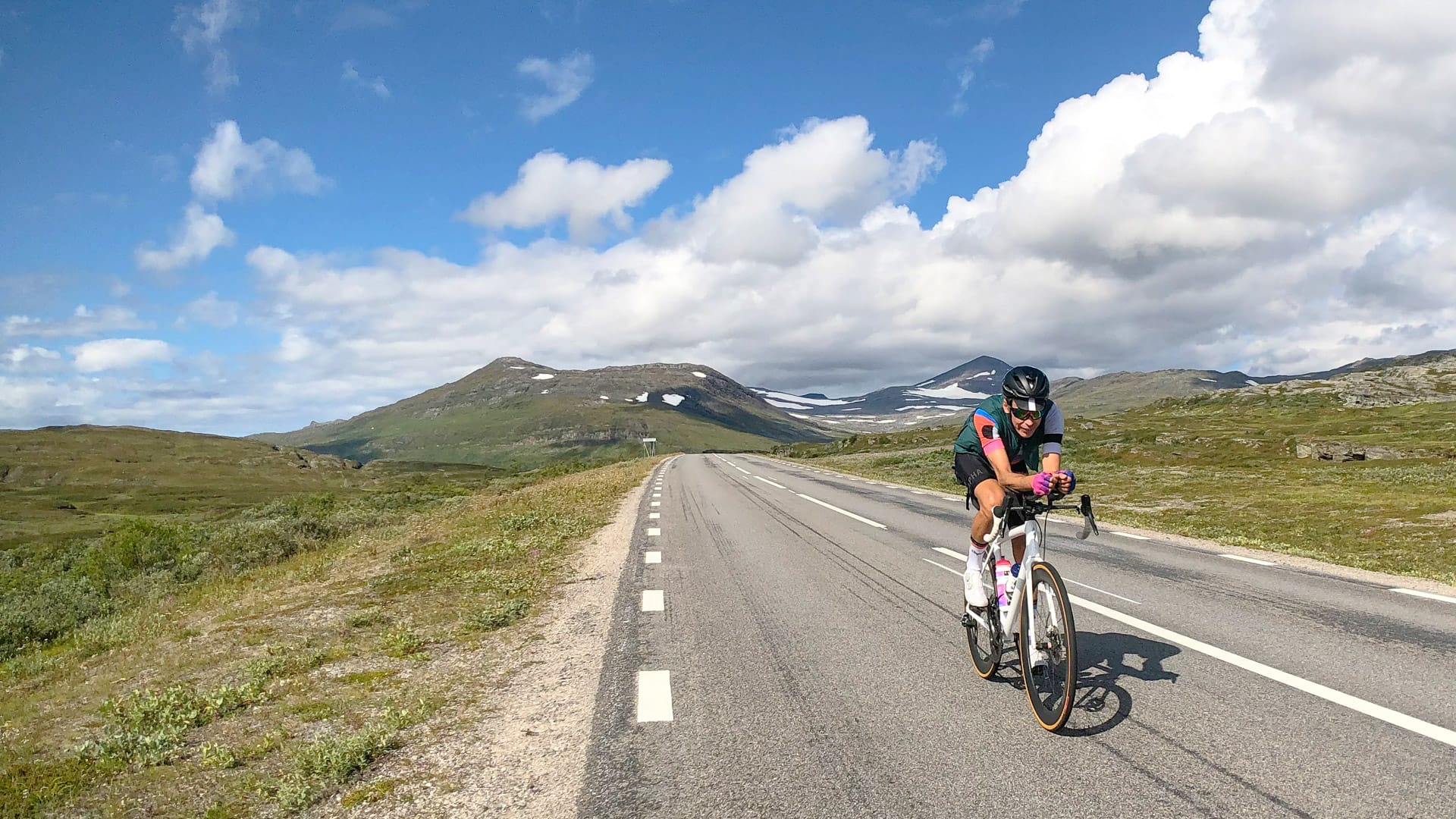 Cyclist riding on a long open road through a mountainous landscape under a partly cloudy sky