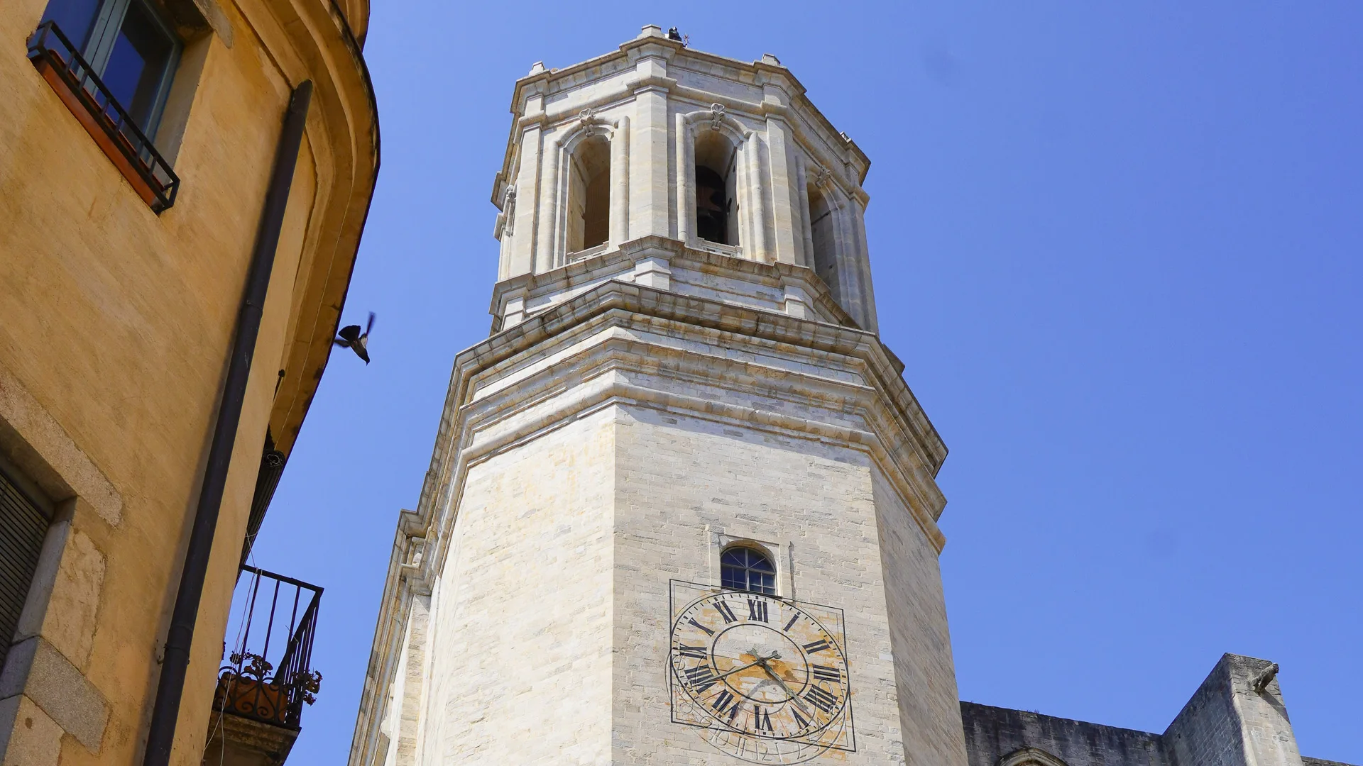 Close-up of Girona Cathedral’s bell tower with clock face and blue sky above narrow street