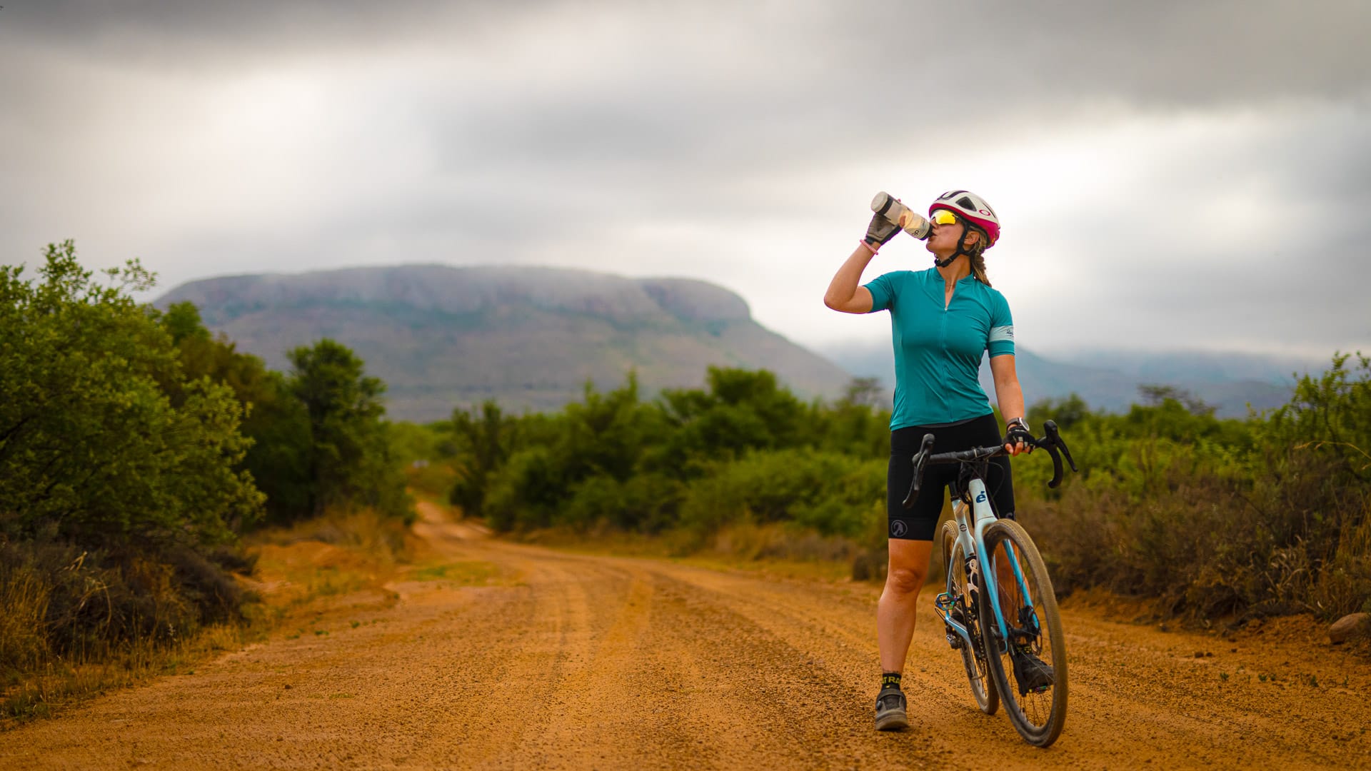 Cyclist standing on a remote dirt road in Africa drinking from a bottle with mountains and moody clouds behind