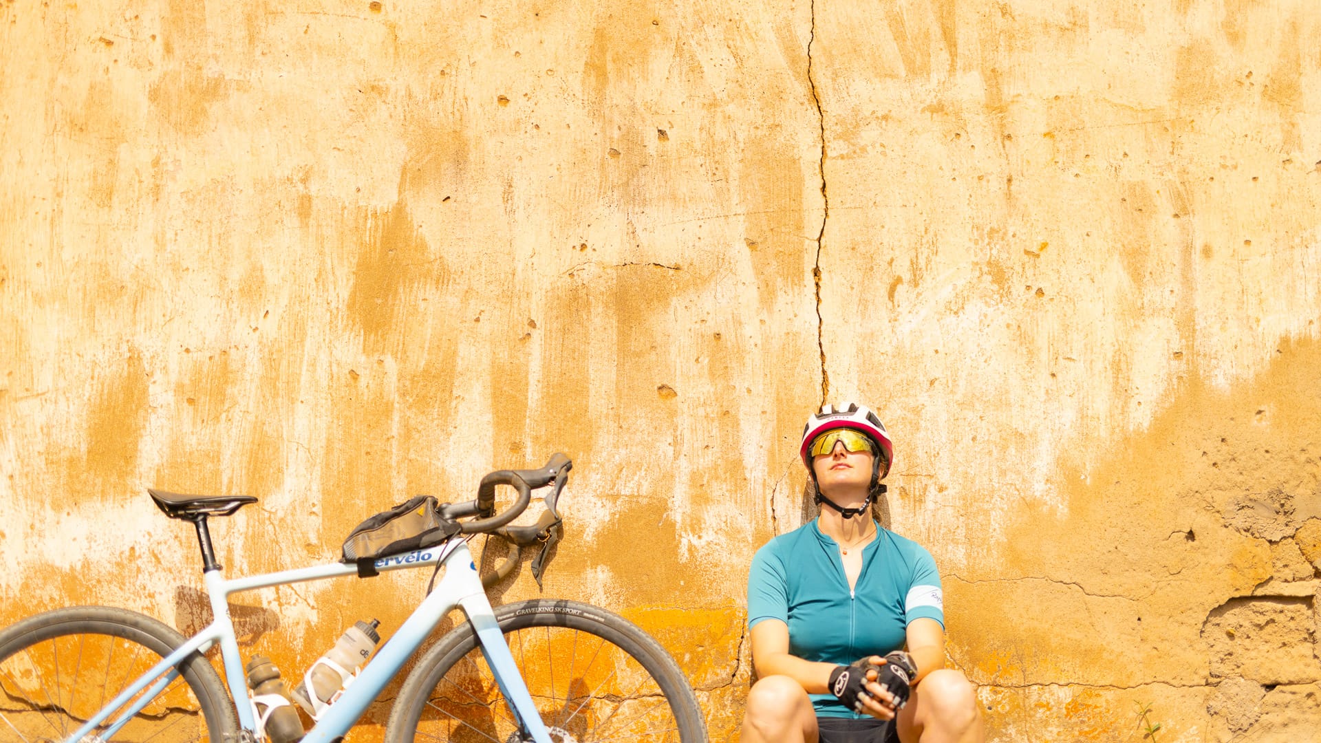 Cyclist resting against a sunlit cracked earth wall with their bike beside them during a hot day in Africa