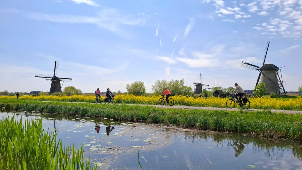 Group of cyclists riding past Dutch windmills on the Amsterdam to Bruges cycling route beside a calm canal