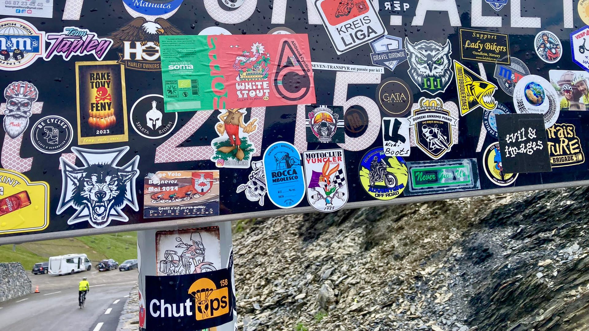 Sticker-covered summit sign on iconic Tour de France climb in the French Alps with cyclist below
